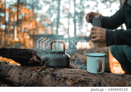 Serene forest camp showing a kettle, an enamel mug, and a person holding a coffee grinder on a log. Warm sunlight filters through the trees, creating a cozy and rustic vibe. Serene forest camp showing a kettle, an enamel mug, and a person holding a coffee grinder on a log. Warm sunlight filters through the trees, creating a cozy and rustic vibe. 140494310