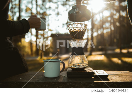 Tranquil outdoor coffee with a kettle pouring water into a pour-over coffee maker on a wooden surface, alongside an enamel mug. Warm sunlight filters through the forest background. 140494424