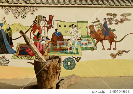 An ancient mortar and pestle by the wall, adorned with paintings depicting life in the Kuche Palace, China. 140494698