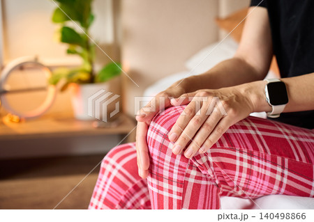 Woman holding painful knee while sitting on bed at home 140498866