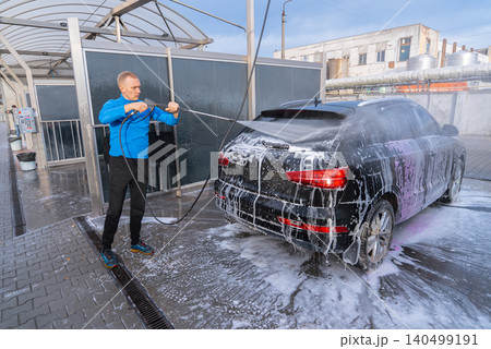 Man Washing Car at Self Service Car Wash Using high pressure Water Cleaning System. Natural daylight highlights the water droplets, foam texture and reflective vehicle surfaces. Concept of vehicle Man Washing Car at Self Service Car Wash Using high pressure Water Cleaning System. Natural daylight highlights the water droplets, foam texture and reflective vehicle surfaces. Concept of vehicle 140499191