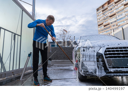 Man Washing Car at Self Service Car Wash. Applying thick foam with a pressure washer. The scene takes place in an outdoor washing bay with modern self-service equipment, automotive accessories Man Washing Car at Self Service Car Wash. Applying thick foam with a pressure washer. The scene takes place in an outdoor washing bay with modern self-service equipment, automotive accessories 140499197