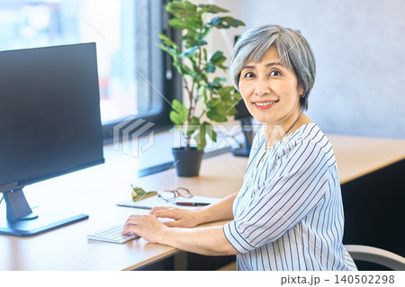 オフィスのパソコン前で笑顔で振り向くグレーヘアの女性 オフィスのパソコン前で笑顔で振り向くグレーヘアの女性 140502298
