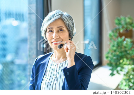 窓辺のオフィスでヘッドセットを付けて微笑むグレーヘアの女性 窓辺のオフィスでヘッドセットを付けて微笑むグレーヘアの女性 140502302