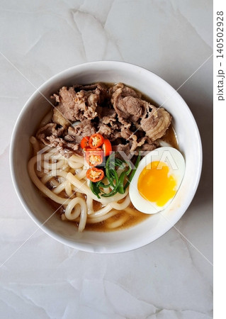 A bowl of udon noodles in a savory broth, topped with green onions, tempura, and a soft boiled egg. The thick, chewy noodles and rich flavors make it a comforting Japanese classic. A bowl of udon noodles in a savory broth, topped with green onions, tempura, and a soft boiled egg. The thick, chewy noodles and rich flavors make it a comforting Japanese classic. 140502928