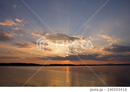 茨城県、涸沼の夕景。オレンジ色に輝く夕日が雲に隠れ、青空に放射状の光差し、水面も輝く。印象的で綺麗だ 140503418
