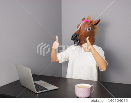 Adult wearing horse head mask giving thumbs up at desk with laptop and coffee mug in studio Adult wearing horse head mask giving thumbs up at desk with laptop and coffee mug in studio 140504036