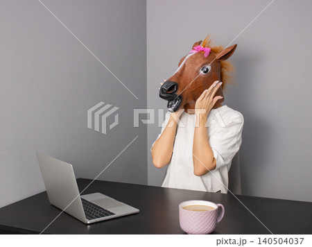 Office worker wearing horse head mask using laptop at desk with coffee cup 140504037