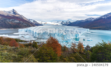 Glacier ice field in mountain lake landscape with snowy peak, autumn forest, and cloudy sky 140504179