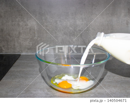 Milk pouring into glass bowl with raw eggs on kitchen counter for homemade batter preparation 140504671