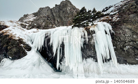 Frozen waterfall and icefall on rocky mountain cliff with deep snow and pine trees in winter Frozen waterfall and icefall on rocky mountain cliff with deep snow and pine trees in winter 140504689