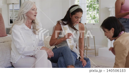 Adjusting seated woman wearing Bride to Be sash touching knee in living room, friends helping Adjusting seated woman wearing Bride to Be sash touching knee in living room, friends helping 140506183
