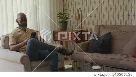 Sitting middle-aged man holding smartphone in armchair in living room, wearing beige shirt Sitting middle-aged man holding smartphone in armchair in living room, wearing beige shirt 140506189