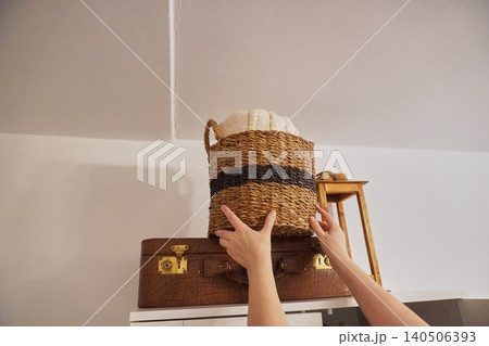 Hands Placing Woven Basket on Top of Vintage Suitcase in Cozy Home Interior Setting 140506393