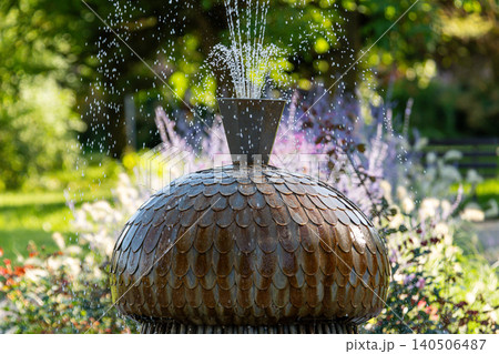 Water fountain releases droplets in a vibrant garden with blooming flowers on a sunny day. 140506487