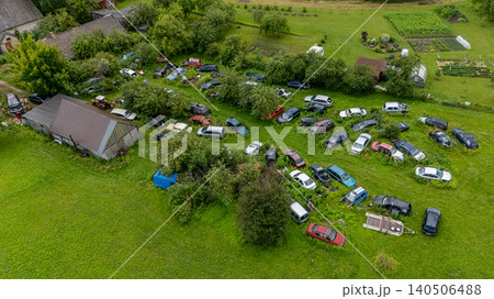 Numerous old cars are abandoned in a grassy area near several rustic buildings and gardens. 140506488