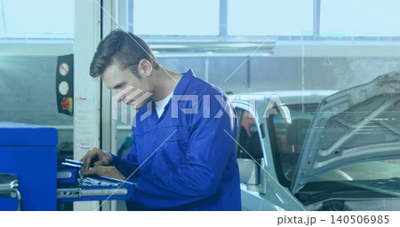 Inspecting mechanic in blue coverall, white tee leaning over tool cart at garage near silver car Inspecting mechanic in blue coverall, white tee leaning over tool cart at garage near silver car 140506985