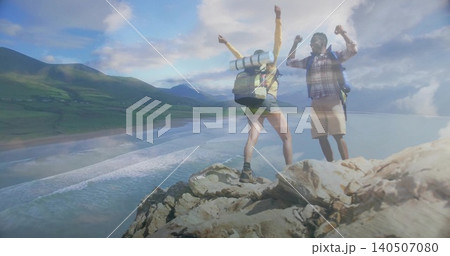Standing hikers raising arms on cliff overlooking sandy bay, in shorts and packs with pad Standing hikers raising arms on cliff overlooking sandy bay, in shorts and packs with pad 140507080