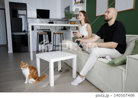Playful young happy couple playing video games on sofa in modern white and green interior at home 140507406