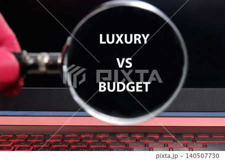 A person holds a magnifying glass in front of a laptop screen displaying the text luxury vs budget. The scene shows decision making in shopping for products A person holds a magnifying glass in front of a laptop screen displaying the text luxury vs budget. The scene shows decision making in shopping for products 140507730