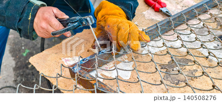 Welder's gloved hands operating an arc welding torch to join metal wire mesh for chain-link fence construction or repair, highlighting skilled, safe industrial craftsmanship. Welder's gloved hands operating an arc welding torch to join metal wire mesh for chain-link fence construction or repair, highlighting skilled, safe industrial craftsmanship. 140508276