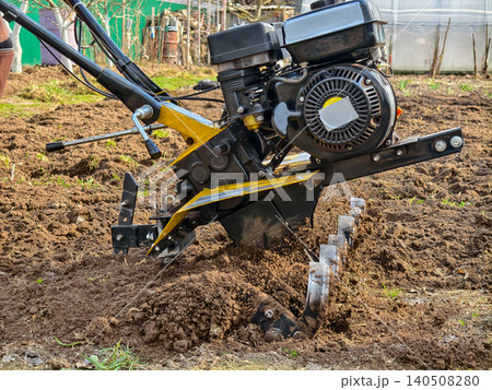 Person using a powerful walk-behind tractor to intensely cultivate freshly turned soil in a garden plot, preparing the ground for planting and seasonal agriculture. Person using a powerful walk-behind tractor to intensely cultivate freshly turned soil in a garden plot, preparing the ground for planting and seasonal agriculture. 140508280