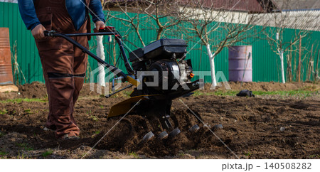 Man cultivating rich soil with a walk-behind tractor in a backyard garden, tilling ground for spring planting on a small farm homestead, preparing beds for growing food. Man cultivating rich soil with a walk-behind tractor in a backyard garden, tilling ground for spring planting on a small farm homestead, preparing beds for growing food. 140508282