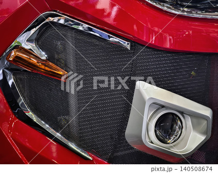 Close-up of a red car front with a grille, fog light, and design details in a modern setting Close-up of a red car front with a grille, fog light, and design details in a modern setting 140508674