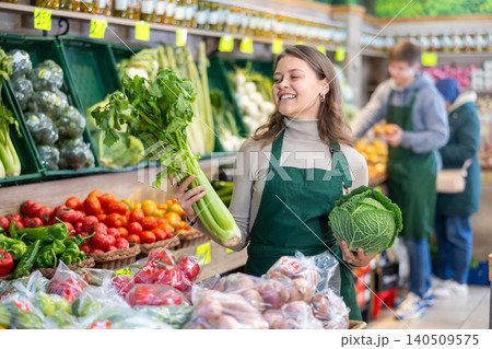 Young female seller holding cabbage and celery standing by counter in grocery market 140509575