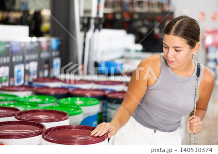 Girl examine bucket of paint, buy collect necessary supplies in store according to list for repairs Girl examine bucket of paint, buy collect necessary supplies in store according to list for repairs 140510374