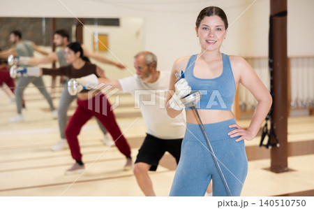 Portrait of sporty woman holding foil in her hands against the background of group of people practicing a foil strike during fencing training in gym Portrait of sporty woman holding foil in her hands against the background of group of people practicing a foil strike during fencing training in gym 140510750