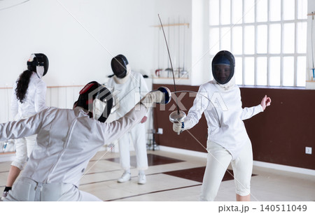 Fencing training - men and women in white uniforms practice fencing in a gymnasium for duel Fencing training - men and women in white uniforms practice fencing in a gymnasium for duel 140511049