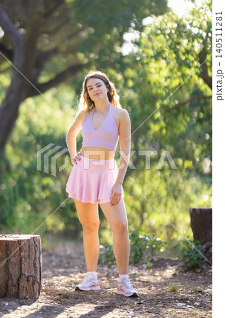 Young woman stands in forest area during daytime while wearing sports attire and posing near tree stump 140511281