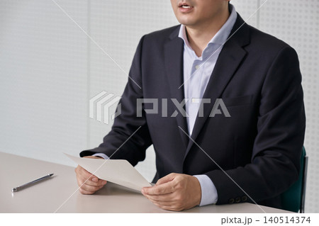 記者会見でスーツ姿の男性が会議室のテーブルで書類を持ち発言や発表を行う顔無しイメージ 140514374