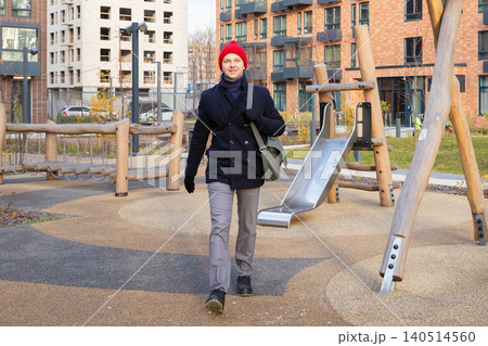 Man walking through modern residential complex courtyard on path to work. Male commuter in casual coat and red hat carrying bag near outdoor playground. Urban lifestyle and commute. Man walking through modern residential complex courtyard on path to work. Male commuter in casual coat and red hat carrying bag near outdoor playground. Urban lifestyle and commute. 140514560