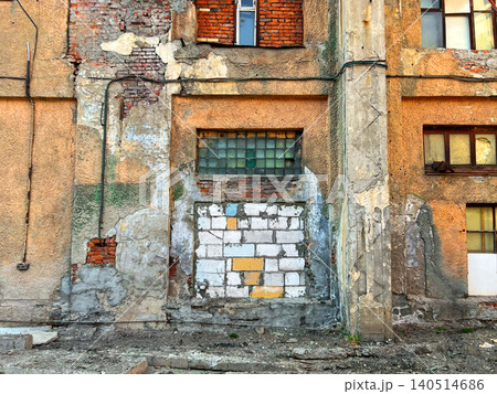 Peeling plaster facade of an abandoned industrial building with old blocked window and exposed brick 140514686