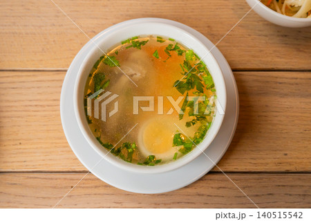 Chicken soup in a white bowl on a wooden table. Homemade broth with sliced egg, carrot and fresh green herbs. Healthy traditional meal served in a rustic dining room setting. Chicken soup in a white bowl on a wooden table. Homemade broth with sliced egg, carrot and fresh green herbs. Healthy traditional meal served in a rustic dining room setting. 140515542