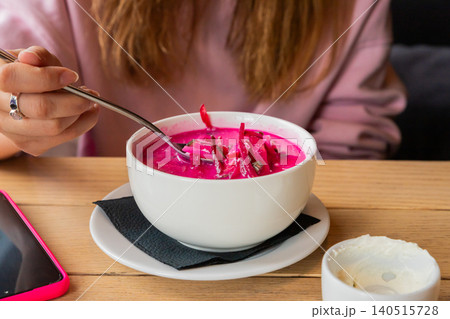 Woman eating traditional Belarusian cold beetroot soup called Kholodnik from white bowl in cafe. Summer refreshing dish with vegetable and herb ingredient for lunch or dinner meal. Woman eating traditional Belarusian cold beetroot soup called Kholodnik from white bowl in cafe. Summer refreshing dish with vegetable and herb ingredient for lunch or dinner meal. 140515728