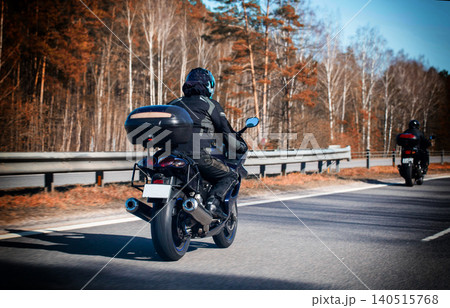 Motorcyclist wearing full protective gear and helmet rides motorcycle with storage box on rural road bordered by pine and birch trees on sunny day journey. 140515768