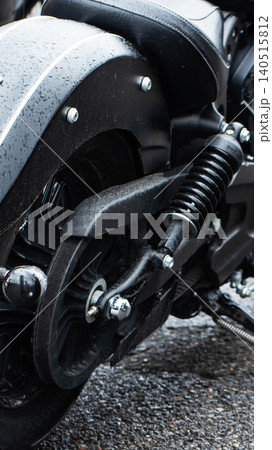 Close-up vertical shot of rear wheel and suspension system of dark cruiser motorcycle, surface details with raindrops, parked outdoors on wet asphalt, mechanical components visible, vertical photo 140515812