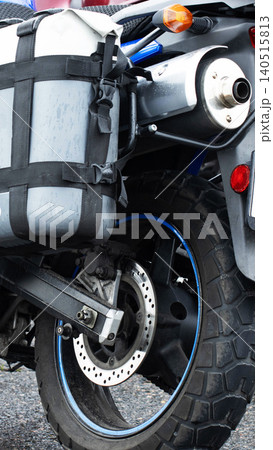 Rear view of adventure motorcycle with knobby tire, brake disk, exhaust system, and waterproof side pannier, featuring wet surface detail and blue rim accent, parked outdoors, vertical photo 140515813
