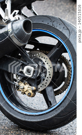 Close-up motorcycle rear wheel wet tire blue rim detail on asphalt. Carbon fiber exhaust pipe, disc brake caliper, drive chain visible rainy conditions, vertical photo Close-up motorcycle rear wheel wet tire blue rim detail on asphalt. Carbon fiber exhaust pipe, disc brake caliper, drive chain visible rainy conditions, vertical photo 140515816