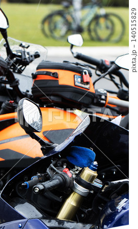 Close-up detail of handlebars, mirrors, and front fairing of two motorcycles parked outdoors during light rain, contrasting blue and orange vehicles with tank bag, vertical composition. Close-up detail of handlebars, mirrors, and front fairing of two motorcycles parked outdoors during light rain, contrasting blue and orange vehicles with tank bag, vertical composition. 140515829