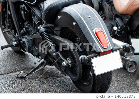 Rear view of modern black motorcycle parked on wet asphalt surface on rainy day. Drops of water visible on fender, taillight, exhaust pipes, and seat highlighting vehicle style. 140515875