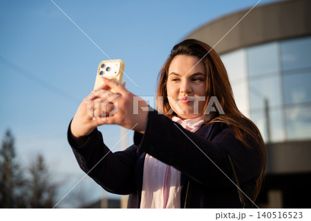 Young businesswoman taking a selfie with a smartphone outside a modern office building in daylight. Digital communication and personal branding for professional online presence Young businesswoman taking a selfie with a smartphone outside a modern office building in daylight. Digital communication and personal branding for professional online presence 140516523