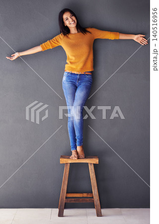 Woman, confident and stool in fashion portrait, smile and style on wall background. Female person, clothing and happy lady on chair, proud and excited in living room at home in house with balance 140516956