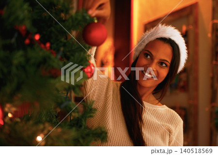 Woman, tree and decoration on Christmas at night, merry celebration and smile for festive holiday. Female person, happy and hat for religious vacation, ornament and prepare for tradition in home Woman, tree and decoration on Christmas at night, merry celebration and smile for festive holiday. Female person, happy and hat for religious vacation, ornament and prepare for tradition in home 140518569
