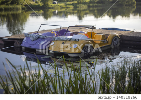 Vintage-style pedal boats shaped like cars moored at a wooden pier on a calm lake Vintage-style pedal boats shaped like cars moored at a wooden pier on a calm lake 140519266
