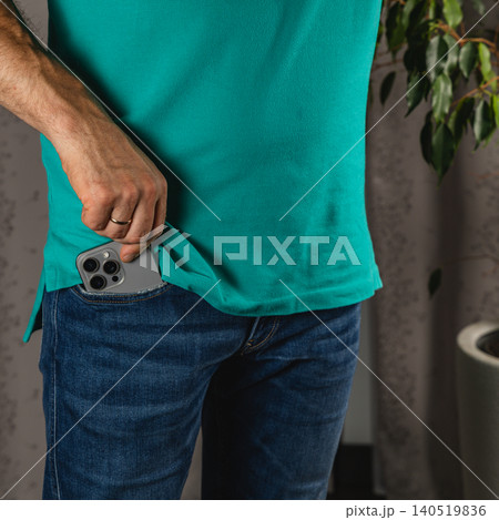 Man puts smartphone into pocket of blue denim jeans while standing indoors Man puts smartphone into pocket of blue denim jeans while standing indoors 140519836