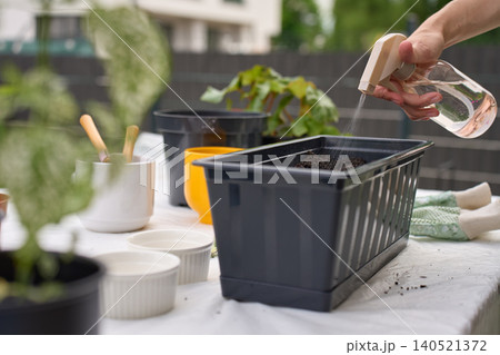 Woman sprays water on soil in planter box at outdoor gardening table Woman sprays water on soil in planter box at outdoor gardening table 140521372